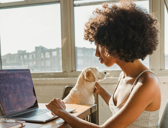 A black woman looking at laptop with a dog sitting on a chair next to her